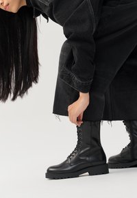 Schwarze Lederkampfstiefel mit strukturierter Oberfläche, runder Zehenpartie und Schnürung, getragen zu einem dunklen Outfit mit fransigen Kanten.