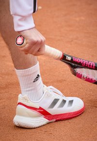 Valkoiset tenniskengät, joissa punaiset yksityiskohdat ja harmaat raidat. Käytetty valkoisen sukan kanssa, jossa musta logo, pitelee tennismaila savipinnalla.