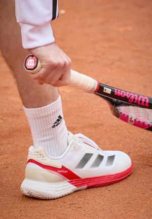 Weiße Tennisschuhe mit roten Akzenten und grauen Streifen. Getragen mit einer weißen Socke, die ein schwarzes Logo zeigt, hält einen Tennisschläger auf einem Sandplatz.
