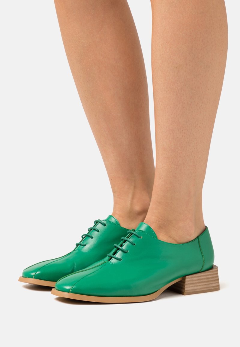 Chaussures en cuir vert à lacets avec petits talons blocs en bois portées pieds nus sur un fond blanc uni.