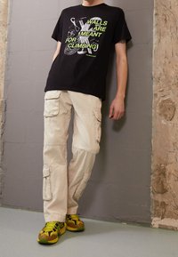 Černé grafické tričko s zeleným nápisem "ZDI JSOU URČENY K LEZENÍ." Béžové cargo kalhoty s kapsami. Žluté a hnědé sportovní boty.