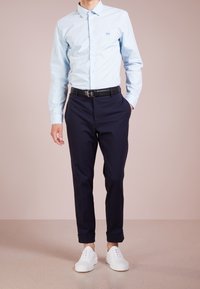 Světle modrá košile na knoflíky, tmavě modré kalhoty a bílé tenisky. Košile má jemné logo; kalhoty jsou na míru, tenisky jsou minimalistické.