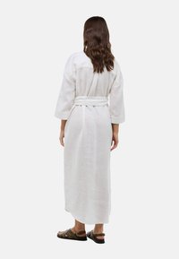 Robe en lin blanc avec des manches longues, ceinturée à la taille, présentant une coupe décontractée et une fente à l'arrière, associée à des sandales marron.