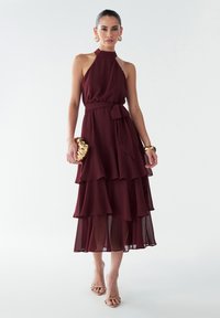 Bordo sluoksniuota suknelė su apykakle, juosmens juosta ir permatomu audiniu; papildyta auksine rankine ir minimaliais sandalais.