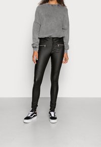 Person som bär svarta läderbyxor med dragkedjefickor, grå långärmad tröja och svart-vita sneakers står på en grått golv.