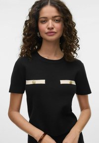 Haut noir à manches courtes avec des accents en tissu blanc et des boutons dorés sur la poitrine. Texture douce et design ajusté.