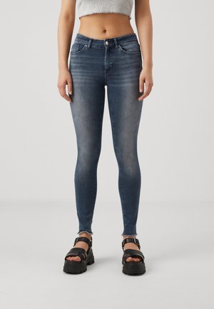 Femme portant un jean skinny bleu délavé, un haut gris court et des sandales à plateforme noires, debout devant un fond clair uni.