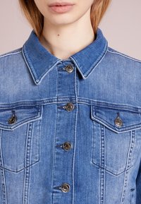 Žena v modré džínové bundě s špičatým límcem a kovovými knoflíky, zobrazená od ramen až po rty na růžovém pozadí.