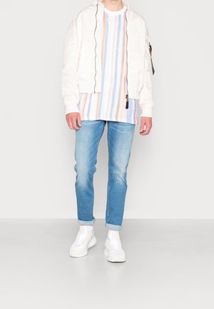 Veste bomber blanche, chemise rayée multicolore, jean bleu clair et baskets blanches. Tenue décontractée avec une coupe ample et des textures douces.