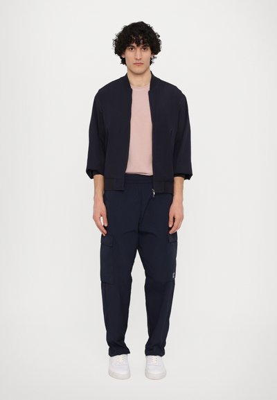 Mornarska bomber jakna z zadrgo, v kombinaciji s svetlo rožnato majico in mornarskimi cargo hlačami z stranskimi žepi, obuta v bele superge.