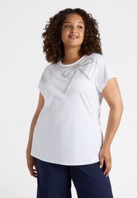 Camiseta corta de manga corta blanca con un lazo plateado decorativo en el escote. Tela suave y ligera con un corte relajado, combinada con pantalones oscuros.