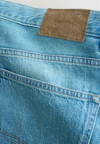 Poche arrière d'un jean en denim bleu clair avec coutures visibles et un patch en cuir marron portant l'inscription "DENIM SUPPLY" sur la ceinture.