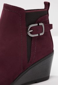 Burgunderfarbene Wildleder-Ankle-Boots mit elastischen Seitenteilen, einem silbernen Schnallenakzent und einem strukturierten schwarzen Keilabsatz.