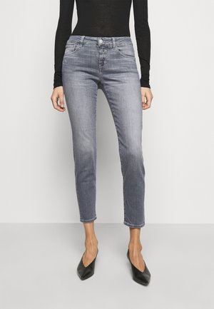 Femme portant un jean skinny gris délavé taille haute, un top noir à manches longues et des talons noirs à bouts pointus, debout devant un fond uni.