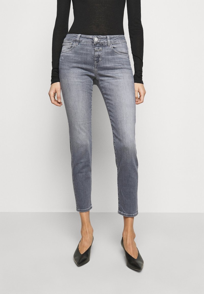 Femme portant un jean skinny gris délavé taille haute, un top noir à manches longues et des talons noirs à bouts pointus, debout devant un fond uni.
