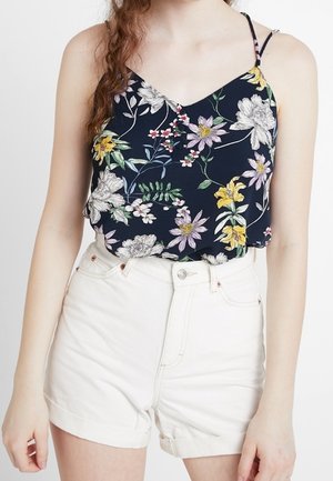 Femme portant un top noir à fines bretelles fleuri, rentré dans un short en jean blanc taille haute sur un fond uni.