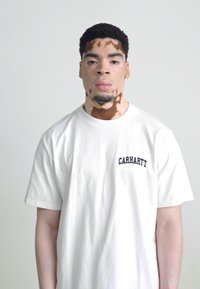 A fehér pamut pólón fekete "CARHARTT" logó található a mellkason. A modellen közepes testalkat és egyedi bőrminta van. Egyszerű háttér.