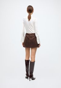 White long-sleeve blouse, brown suede mini skirt, and glossy knee-high boots with a stiletto heel, facing away from the camera.