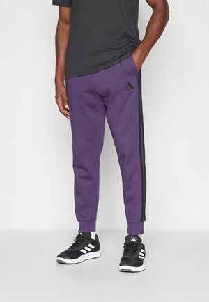 Lilla joggebukser med svarte sidelister, elastiske mansjetter, og et brodert svart Adidas-logo på venstre lår. Kombinert med svarte treningssko.
