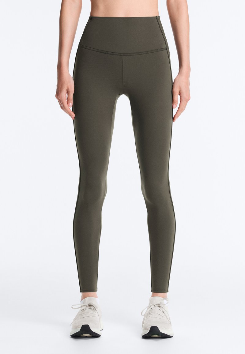 Legging in olijfgroen met een hoge taille en een nauwsluitende pasvorm, gedragen met witte sportschoenen door een staand persoon tegen een effen achtergrond.