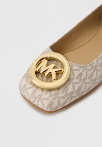 Chaussure plate beige avec un motif texturé, ornée d'un logo circulaire doré proéminent. Design à bout carré, intérieur lisse, et accents subtils.