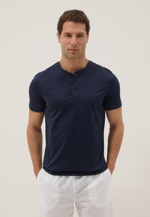 Mann trägt ein marineblaues Henley-Shirt mit kurzen Ärmeln und weiße Hose, steht mit den Händen in den Taschen vor einem schlichten Hintergrund.