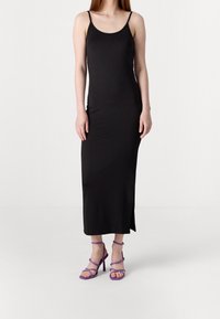 Černé, bezrukávové maxi šaty s přiléhavým střihem a bočním rozparkem; spárováno s fialovými sandály na vysokém podpatku se stahovacími pásky.