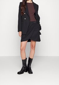 Svart skräddarsydd kavaj med guldknappar, brunt ribbat linne, svart draperad kjol och svarta läderstövlar med klack. Släta texturer och modern silhuett.