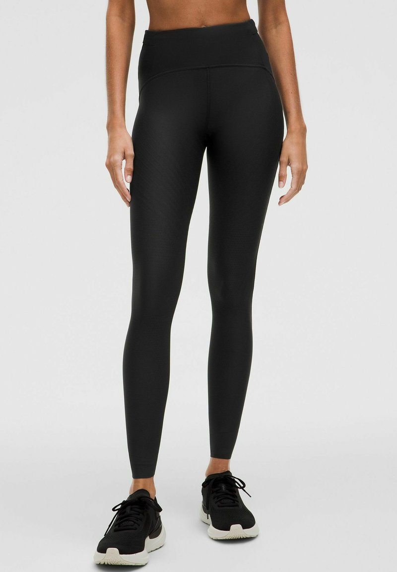 Schwarze hochtaillierte Leggings mit glatter Textur, ausgestattet mit einem dezenten diagonalen Muster und einer figurbetonten Passform, kombiniert mit schwarzen Sportschuhen.