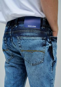 Vaqueros de mezclilla azul de cintura alta con un lavado deslavado, costuras doradas y dos bolsillos traseros. Las etiquetas visibles en la cintura dicen "SALSA JEANS."