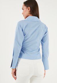 Donna con lunghi capelli castani che indossa orecchini a cerchio dorati, una camicia a righe blu e bianche a maniche lunghe e pantaloni bianchi, di spalle.