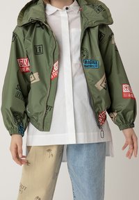 Giacca verde leggera con chiusura a zip, polsini elastici e vari toppe stampate colorate. Abbinata a pantaloni chiari di diversi colori.