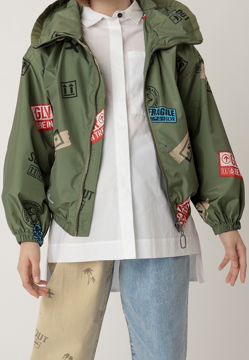 Giacca verde leggera con chiusura a zip, polsini elastici e vari toppe stampate colorate. Abbinata a pantaloni chiari di diversi colori.