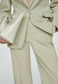 Vrouw in een beige op maat gemaakt pak die met één hand een lichtbeige leren handtas vasthoudt terwijl ze met de andere hand in haar zak zit.