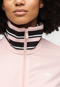 Ροζ ζακέτα με φερμουάρ, που διαθέτει ψηλό γιακά με μαύρες και λευκές ρίγες, λείο ύφασμα και μικρό λευκό λογότυπο στην αριστερή πλευρά.