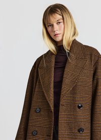 Brauner karierter Doppelreiher mit großen Revers, ausgestattet mit schwarzen Knöpfen und einem strukturierten Stoff. Getragen über einem dunklen Rollkragenpullover.