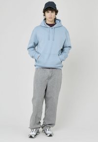 Haut à capuche bleu clair avec poche kangourou, cordons au niveau du col, associé à un pantalon large gris clair et des chaussures de sport blanches.