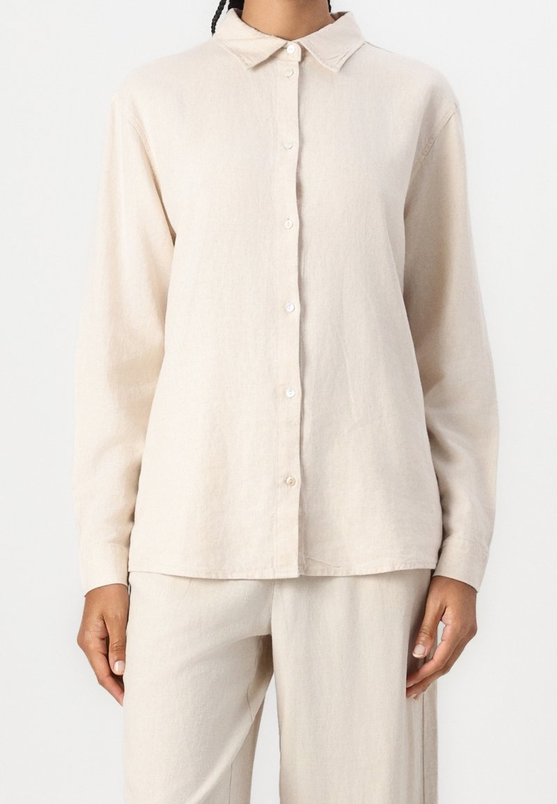 Chemise en lin beige avec un devant à boutons, des manches longues, un col et une coupe décontractée. Le tissu a une apparence texturée.