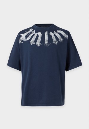 Άνδρας που φοράει ένα μπλε σκούρο oversized T-shirt με λευκό κείμενο και φαρδιά γκρι παντελόνια, στέκεται με τα χέρια στις τσέπες σε μονόχρωμο φόντο.