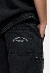Pantalons cargo en denim noir avec une poche latérale, un logo brodé sur la poche arrière et une coupe décontractée. Tissu texturé et matériel robuste.