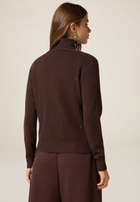 Suéter marrón de punto con cuello alto, dobladillo y puños acanalados, adornado con sutiles motas. La espalda del modelo está de espaldas a la cámara.
