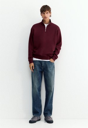 Jeune homme portant un pull bordeaux à demi-fermeture éclair, un t-shirt blanc, un jean bleu et des baskets grises, se tenant devant un fond blanc uni.