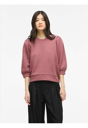 Ung kvinde med langt mørkt hår iført en lavendelfarvet sweatshirt med puffede ærmer og sorte vidde plisserede bukser, står foran en hvid baggrund.