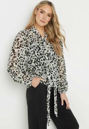 Blouse légère et transparente avec un motif à pois noir et blanc, des manches bouffantes longues et un col à nouer, associée à un pantalon noir.