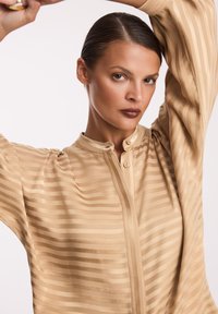 Beżowa bluzka z pionowymi paskami, stójką i zapięciem na guziki z przodu. Materiał ma gładką teksturę, lekki połysk oraz bufiaste rękawy.
