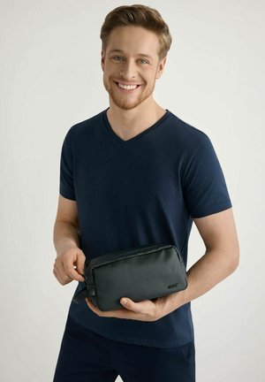 Młody mężczyzna w granatowej koszuli uśmiecha się, trzymając i częściowo rozpinając małą czarną prostokątną saszetkę na jednolitym tle.