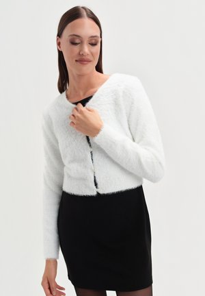 Kobieta w białym, puszystym, krótkim kardiganie zapinanym na perłowe guziki, nosząca czarną sukienkę, stojąca na tle jednolitego białego tła.