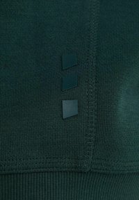 Tessuto di felpa verde scuro con una finitura a maglia testurizzata, caratterizzato da tre piccoli accenti quadrati scuri su un lato e un orlo a costine.