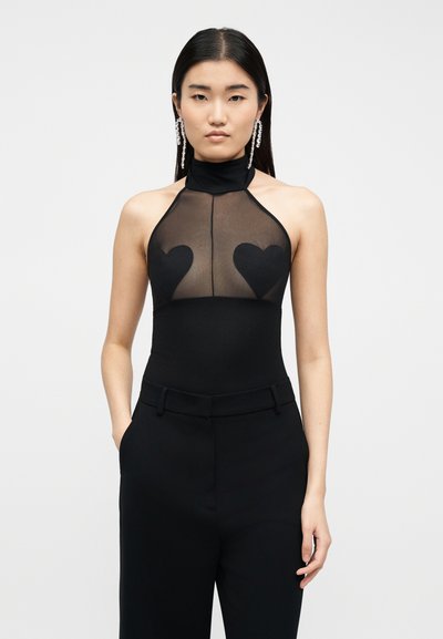 Femme aux longs cheveux noirs portant un pantalon noir taille haute, un haut noir sans manches avec un panneau transparent et des patchs en forme de cœur, ainsi que de longues boucles d'oreilles en cristal.