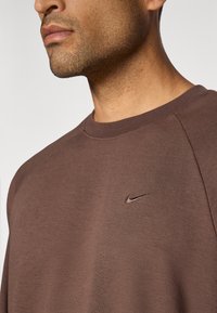 Hnedá mikina s okrúhlym výstrihom, s jemne vyšitým logom Nike na hrudi. Hladká textúra, neformálny dizajn a priliehavé rukávy.